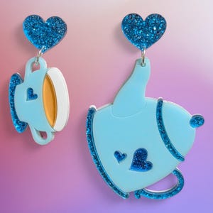 May include: Light blue acrylic earrings featuring a teacup and teapot design. Each earring has a glittery blue heart at the top. The teacup has a white rim and orange interior. The teapot has a small blue heart detail.