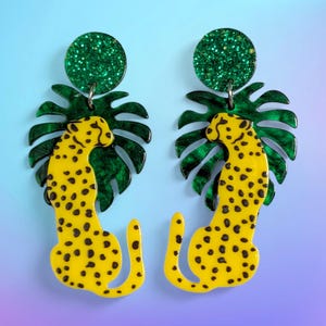 Puede incluir: Un par de pendientes con un diseño de leopardo amarillo con manchas negras. El leopardo está sobre una hoja de monstera verde y un círculo verde brillante. Un accesorio llamativo y audaz.