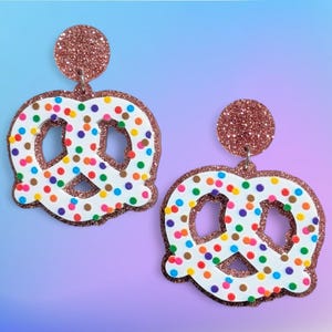 Op de afbeelding: Een paar novelty oorbellen in de vorm van een pretzel. De oorbellen hebben een witte basis met kleurrijke sprinkles en een glinsterende koperkleurige omtrek en bovenste cirkel. De achtergrond is een verloop van blauw en paars.