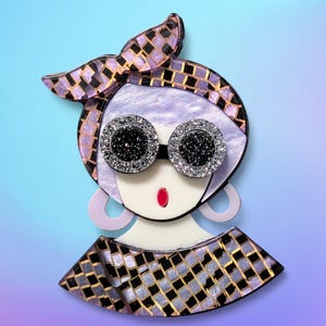 May include: A brooch depicting a stylized face with large, sparkling sunglasses. The design incorporates a headscarf and a geometric pattern in black, gold, and purple. The brooch features a red lip and large, light purple hoop earrings.