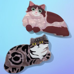 Puede incluir: Dos broches decorativos con forma de gato. El broche superior presenta un gato marrón y blanco con detalles en rosa. El broche inferior representa un gato atigrado gris y negro con ojos amarillos. Ambos broches están sobre un fondo azul degradado.