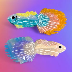 May include: Two colorful fish-shaped hair clips. One clip is blue, yellow, and orange, the other is blue and beige. Both have white accents and black eyes. The clips are on a gradient purple and pink background.