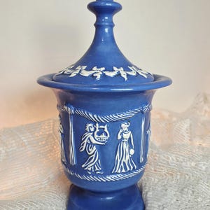 May include: A blue ceramic jar with a matching lid, featuring white relief figures of people and draped garlands. The vessel has a flared rim, a tapered body, and a pedestal base, with a pointed finial on top of the lid.