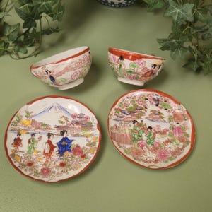 May include: Four pieces of Japanese porcelain tableware. Two small bowls and two plates with a red trim. The plates feature scenes with figures, flowers, and a mountain. The bowls have a similar design. The pieces are on a green surface.