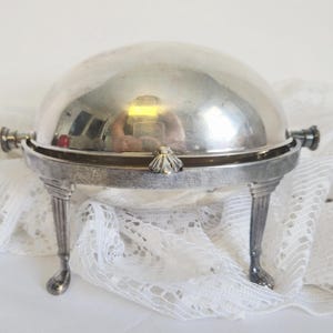 May include: A silver-colored, domed serving dish with a hinged lid. The dish has a decorative, ornate design with a small, shell-shaped handle on the front and two handles on the sides. It stands on four decorative legs.