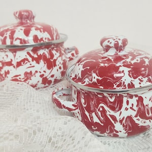 May include: Two red and white enamelware pots with lids. The pots have a speckled pattern and silver rims and handles. The pots are sitting on a white lace surface. The pots are a classic kitchenware item.