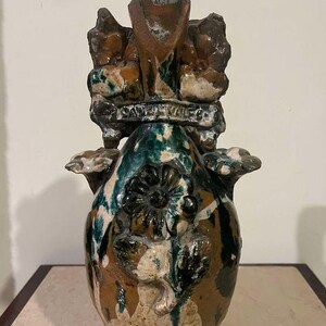 Puede incluir: Un jarrón de cerámica con un diseño único. El jarrón presenta una combinación de colores marrón, verde y blanco con un patrón floral y el texto "SANTA VALERIA". El jarrón tiene una superficie texturizada y una forma irregular, lo que aumenta su atractivo artístico.