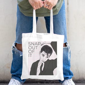 以下が含まれることがあります： ピンクと白のグリッドの背景に「SNAP OUT OF IT」というテキストと、白黒の男性のイラストが描かれた白いトートバッグです。