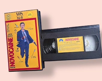 Könnte beinhalten: Ein VHS-Kassetten-Etui mit gelbem Hintergrund und einem Schwarzweißbild eines Mannes, der rennt. Der Titel des Films ist "Novocain" und der Name des Schauspielers ist "Jack Quaid".