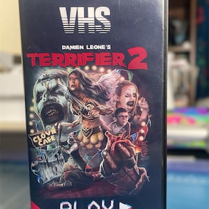 May include: VHS tape cover for the horror movie "Damien Leone's Terrifier 2". The cover features a dark, bloody, and chaotic scene with multiple characters, including a clown, a woman in a warrior costume, and a man with a bloody heart. The text "PLAY" is in a pixelated style with a play button icon.