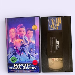 May include: A VHS tape and case for "KPOP Demon Hunters." The case features a colorful illustration of animated characters. The VHS tape is black with a label that reads "KPOP Demon Hunters."