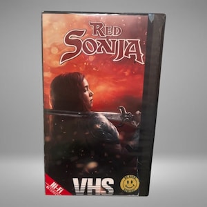 May include: A VHS tape of the movie "Red Sonja" featuring a woman with long hair holding a sword. The title is in red, with a fiery background. The VHS tape has "VHS" and "Hi-Fi Stereo" labels.
