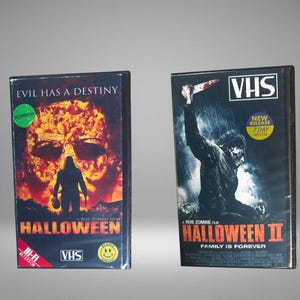 May include: Two VHS tapes of the horror movie "Halloween" and "Halloween II". The first tape has a skull image and the word "Halloween" in red. The second tape shows a man holding a knife in the rain.
