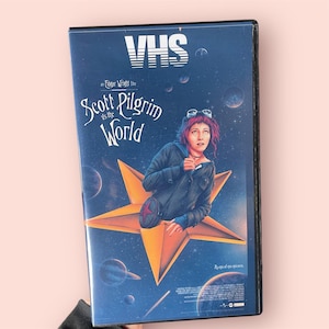 May include: A VHS tape case for the film Scott Pilgrim vs. the World. The cover features a dark blue space background with planets, a large orange star, and a person with pink hair wearing a dark jacket. The text reads VHS, an Edgar Wright film, Scott Pilgrim vs. the World, and An epic of epic epicness.