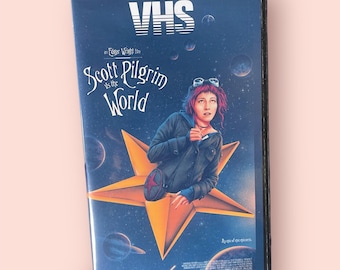Könnte beinhalten: Eine VHS-Kassette des Films Scott Pilgrim vs. the World. Das Cover zeigt einen dunkelblauen Weltraumhintergrund mit Planeten, einem großen orangefarbenen Stern und einer Person mit rosa Haaren in einer dunklen Jacke. Der Text lautet VHS, an Edgar Wright film, Scott Pilgrim vs. the World und An epic of epic epicness.