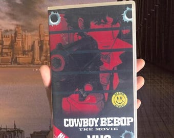 May include: A Cowboy Bebop The Movie VHS tape with a red and black cover featuring a character holding a firearm. The cover includes bullet hole graphics, a yellow smiley face sticker with the text Be Kind Rewind, and a red Hi-Fi Stereo label.