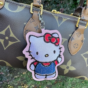 Pode incluir: Um chaveiro Hello Kitty rosa e branco com um laço vermelho e uma roupa em estilo jeans azul. O chaveiro está preso a uma bolsa de grife marrom e bege com detalhes dourados. O fundo é folhagem verde desfocada.