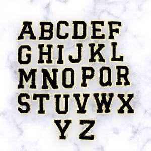 Puede incluir: Letras del alfabeto de fieltro negro con contorno dorado. Las letras están dispuestas en orden alfabético en tres filas.