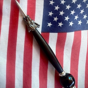 May include: A pointed metal tool featuring a silver-toned wolf head accent at the top, a smooth black cylindrical handle, and a decorative base consisting of a large black spherical bead flanked by two smaller silver-toned textured metal spacers.