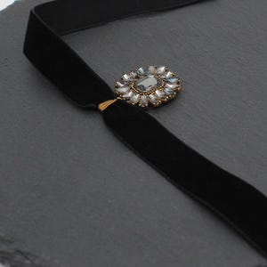 May include: A black velvet choker necklace with a decorative centerpiece. The centerpiece is a gold-toned setting with a large, gray-colored stone surrounded by smaller, clear stones. The necklace is displayed on a dark gray surface.