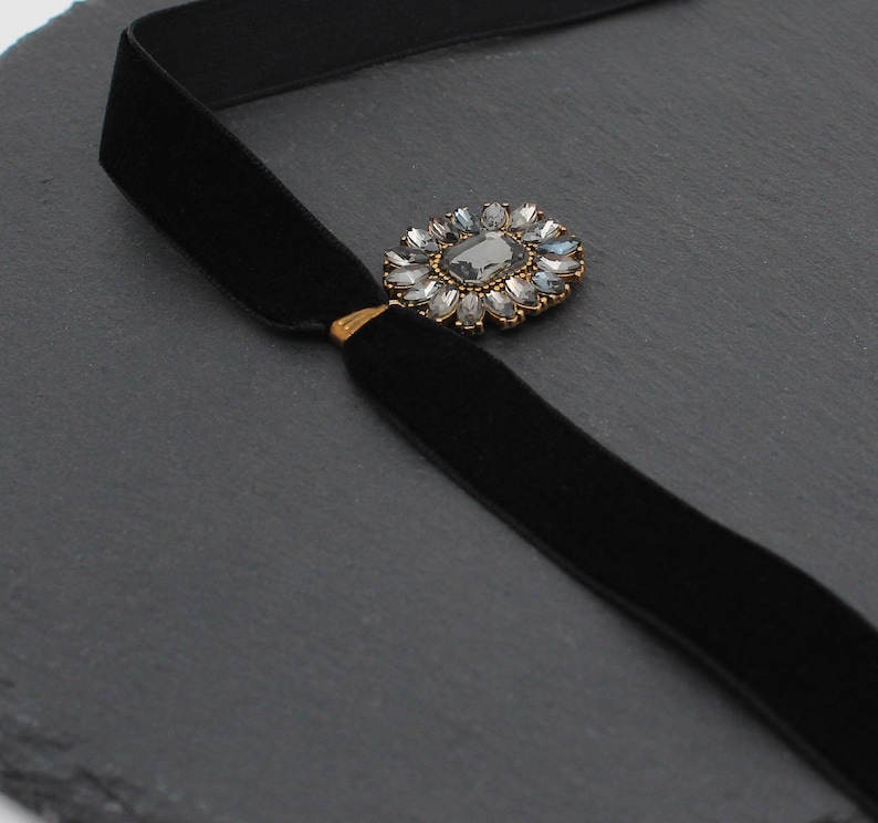 May include: A black velvet choker necklace with a decorative centerpiece. The centerpiece is a gold-toned setting with a large, gray-colored stone surrounded by smaller, clear stones. The necklace is displayed on a dark gray surface.