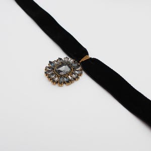 May include: A black velvet choker necklace with a gold-toned floral pendant. The pendant features a large, gray, rectangular center stone surrounded by smaller, gray, teardrop-shaped stones. The necklace is displayed on a white background.