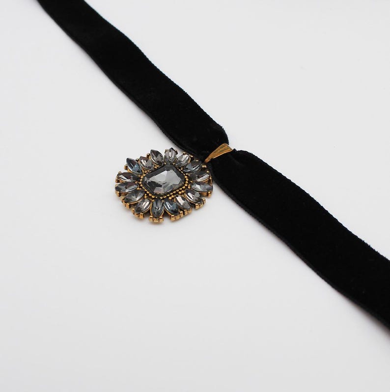May include: A black velvet choker necklace with a gold-toned floral pendant. The pendant features a large, gray, rectangular center stone surrounded by smaller, gray, teardrop-shaped stones. The necklace is displayed on a white background.