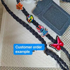 May include: A dark blue braided lanyard featuring a denim-textured ID badge holder and several charms, including an orange maple leaf, a colorful Rubik's cube, a blue alien character, and a pink butterfly. Text reads: Customer order example.