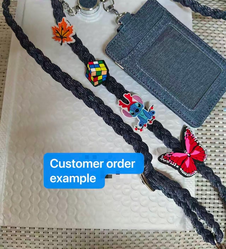 May include: A dark blue braided lanyard featuring a denim-textured ID badge holder and several charms, including an orange maple leaf, a colorful Rubik's cube, a blue alien character, and a pink butterfly. Text reads: Customer order example.