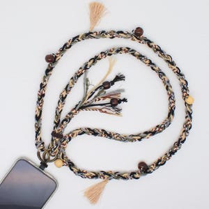 May include: A braided phone lanyard featuring a multi-colored cord in black, white, grey, and tan. The strap is accented with dark brown and light wooden beads, finished with beige tassels at the ends and a metal ring attachment for a mobile device.