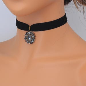 May include: Black velvet choker necklace with a pendant. The pendant features a central square-cut gray stone surrounded by smaller gray stones in a floral design, set in a gold-toned metal frame. The necklace is a classic accessory.