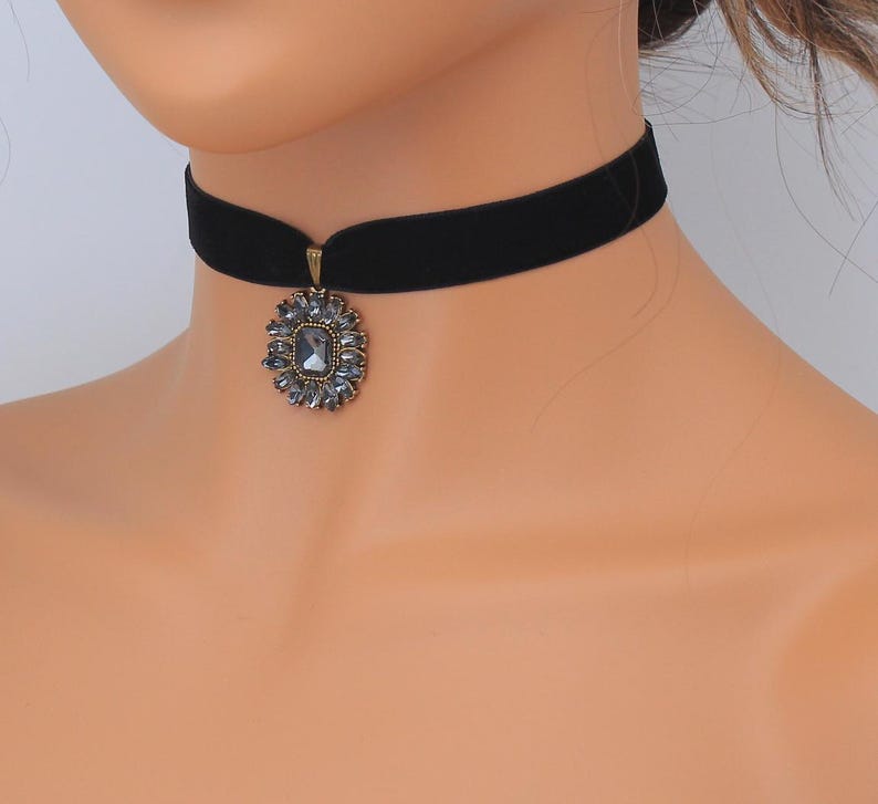 May include: Black velvet choker necklace with a pendant. The pendant features a central square-cut gray stone surrounded by smaller gray stones in a floral design, set in a gold-toned metal frame. The necklace is a classic accessory.