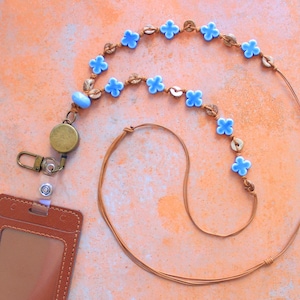 May include: A tan cord lanyard featuring light blue four-petaled flower beads and small round textured brown beads. The lanyard includes a circular bronze-toned retractable badge reel with a metal lobster clasp and a brown rectangular ID holder.