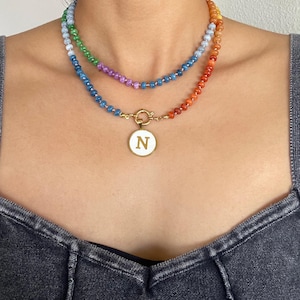Puede incluir: Un collar de varias capas con dos hileras de peque&ntilde;as cuentas multicolores en tonos azul, verde, morado y naranja. La hilera inferior incluye un colgante circular dorado con un centro blanco y la letra N en color dorado.