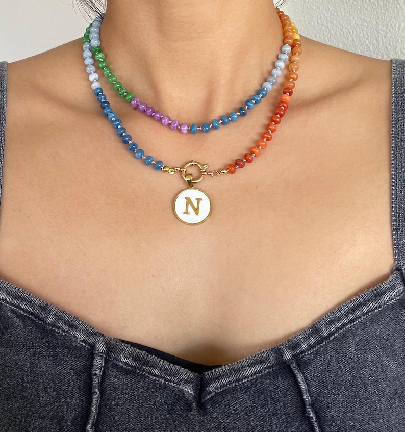 Puede incluir: Un collar de varias capas con dos hileras de peque&ntilde;as cuentas multicolores en tonos azul, verde, morado y naranja. La hilera inferior incluye un colgante circular dorado con un centro blanco y la letra N en color dorado.