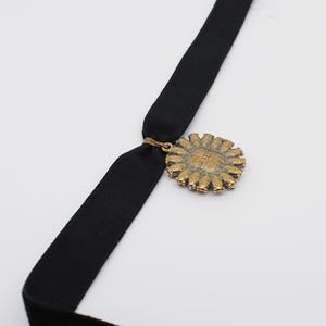 May include: A black velvet choker necklace with a gold-toned pendant. The pendant is circular with a detailed, sunburst design. The necklace is displayed against a white background.