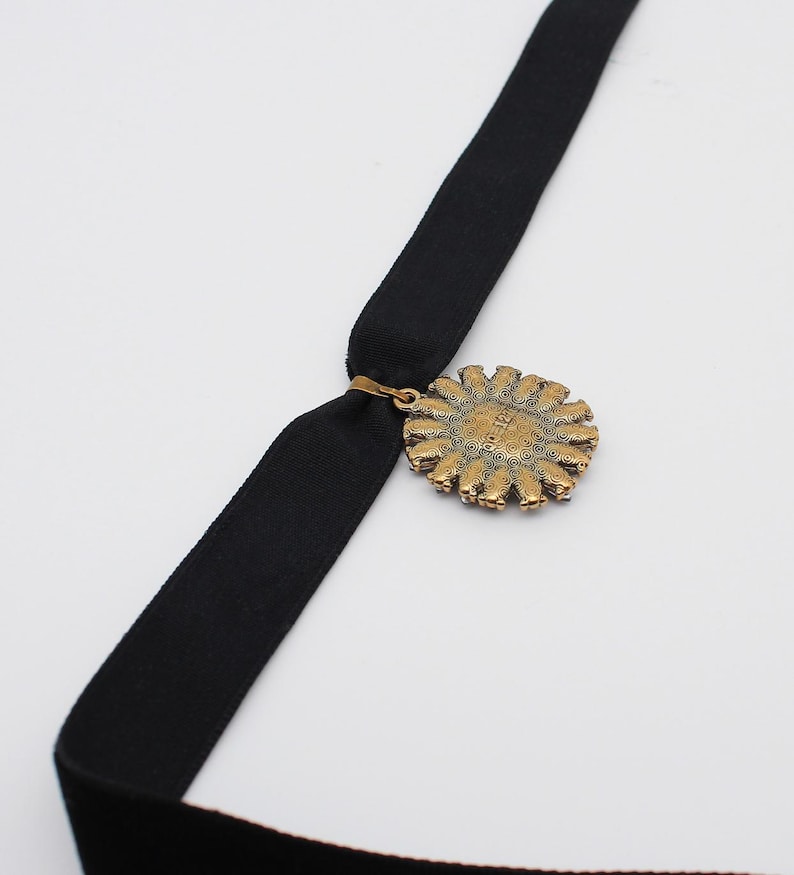 May include: A black velvet choker necklace with a gold-toned pendant. The pendant is circular with a detailed, sunburst design. The necklace is displayed against a white background.