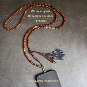 May include: A multi-layer necklace with a phone lanyard, also usable as a bracelet. The necklace is made of small, round, reddish-brown beads. It features a leather tassel with blue and brown accents. The phone is attached with a silver-colored clasp. Text on the image: "Phone lanyard, Multi-layer necklace / bracelet, Tiger Creek Handmade."