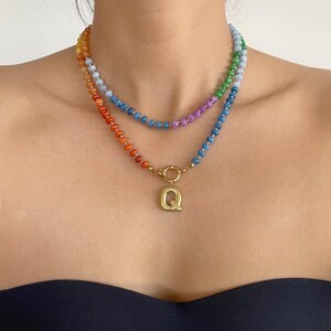 Puede incluir: Un collar de dos capas con peque&ntilde;as cuentas multicolores en tonos naranja, rojo, azul, verde y morado. La capa inferior incluye un cierre circular de tono dorado y un llamativo colgante con la letra Q en tono dorado.