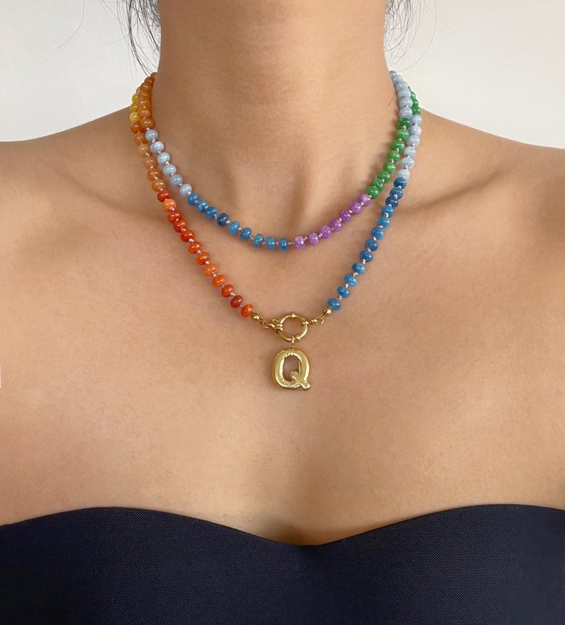 Puede incluir: Un collar de dos capas con peque&ntilde;as cuentas multicolores en tonos naranja, rojo, azul, verde y morado. La capa inferior incluye un cierre circular de tono dorado y un llamativo colgante con la letra Q en tono dorado.