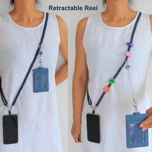 May include: A dark blue braided lanyard featuring a retractable reel, a denim-textured ID badge holder with a clear window, and a black smartphone case. The lanyard is adorned with small colorful decorative pins and charms.