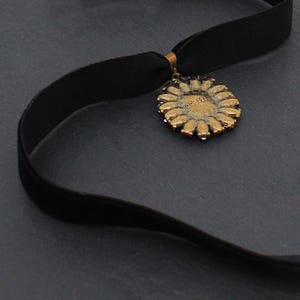 May include: A black velvet choker necklace with a gold-colored pendant. The pendant is circular with a floral design and the text "00 12" in the center. The necklace is displayed on a dark gray surface.