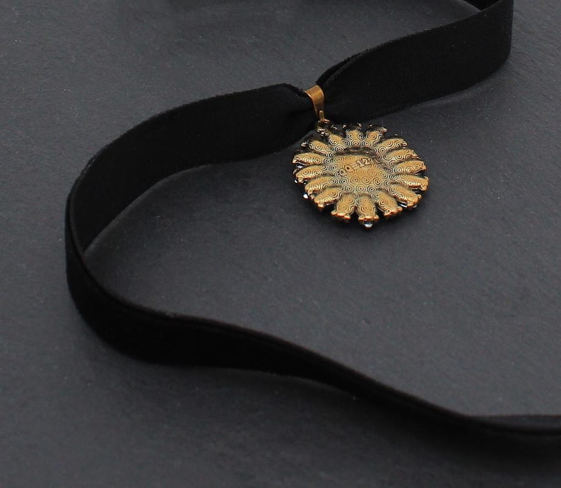 May include: A black velvet choker necklace with a gold-colored pendant. The pendant is circular with a floral design and the text "00 12" in the center. The necklace is displayed on a dark gray surface.
