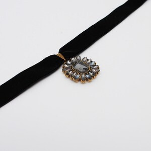 May include: A black velvet choker necklace with a gold-toned clasp and a decorative pendant. The pendant features a large, gray rectangular stone surrounded by smaller, clear and gray stones in a floral arrangement.