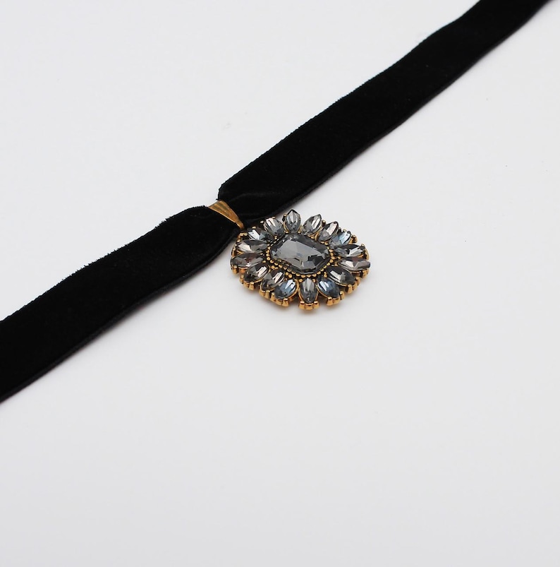 May include: A black velvet choker necklace with a gold-toned clasp and a decorative pendant. The pendant features a large, gray rectangular stone surrounded by smaller, clear and gray stones in a floral arrangement.