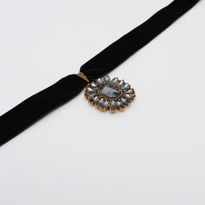 May include: A black velvet choker necklace with a gold-toned clasp and a decorative pendant. The pendant features a large, gray rectangular stone surrounded by smaller, blue-gray stones in a floral arrangement. The necklace is set against a white background.