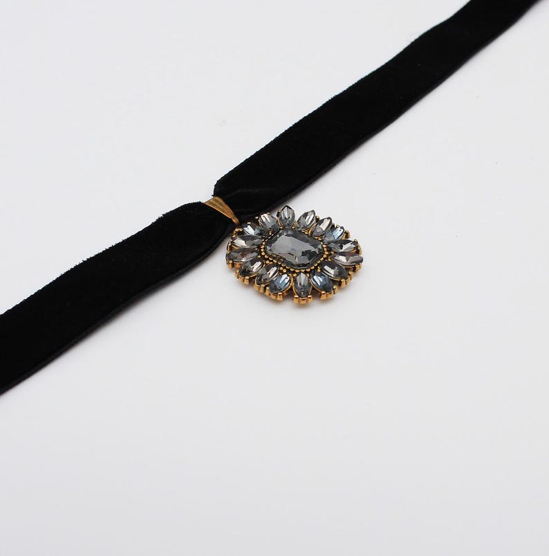 May include: A black velvet choker necklace with a gold-toned clasp and a decorative pendant. The pendant features a large, gray rectangular stone surrounded by smaller, blue-gray stones in a floral arrangement. The necklace is set against a white background.