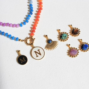Puede incluir: Un collar de cuentas multicolor con un cierre dorado y dos colgantes circulares con las letras M y N, junto a cuatro colgantes dorados adicionales con piedras de varios colores y un dise&ntilde;o de rayos de sol.