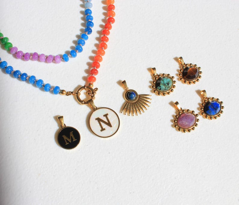 Puede incluir: Un collar de cuentas multicolor con un cierre dorado y dos colgantes circulares con las letras M y N, junto a cuatro colgantes dorados adicionales con piedras de varios colores y un dise&ntilde;o de rayos de sol.
