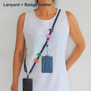 May include: A dark blue braided lanyard featuring multiple colorful pins, a retractable badge reel, and a denim-textured badge holder. The lanyard includes a metal clip for a phone and the text Lanyard + Badge holder is visible.